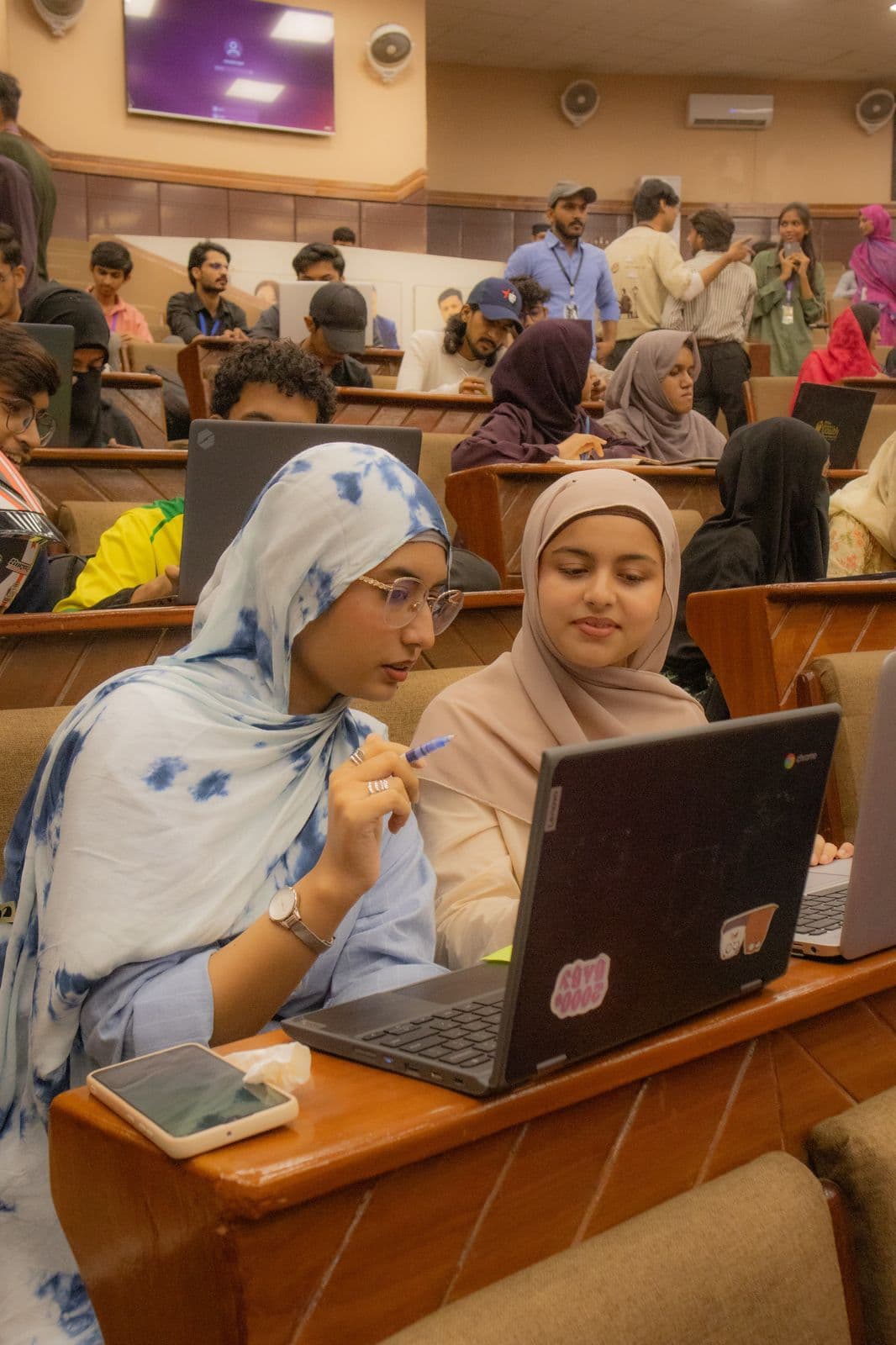 Two attendees sharing a laptop in the front row of a university lecture hall at Session 6, with the rest of the hall filling up behind them